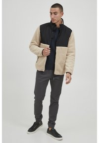 Fleece jacket with black upper section and beige lower section, featuring a high collar, zip closure, and side pockets. Worn with dark pants and sneakers.