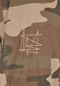 Maskovacia látka v zemských odtieňoch má abstraktný prešitý dizajn v svetlejšej niťi, čo zvýrazňuje textúru a vzor.