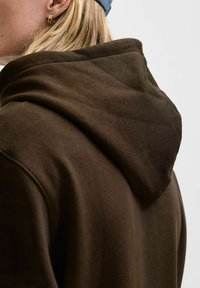 Brown hooded sweatshirt features a soft texture, rounded hood, and stitched seams, worn with a blue cap, highlighting the garment's design.