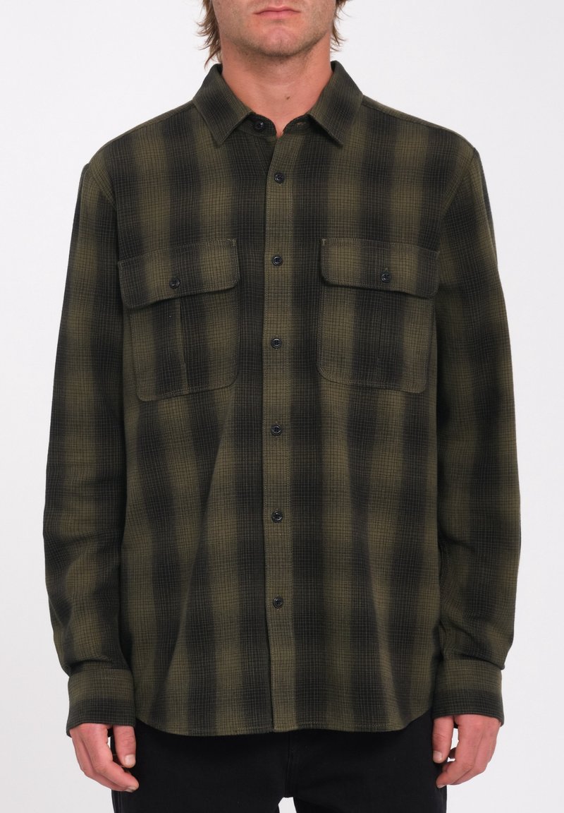 Homme portant une chemise à manches longues à boutons, à carreaux verts et noirs, avec deux poches poitrine sur le devant, se tenant devant un fond uni.