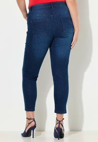 Tmavě modré džíny z denimu s přiléhavým střihem a dvěma zadními kapsami. Okraj nohavic je zatočený, odhalující sandály na vysokém podpatku s pásky.