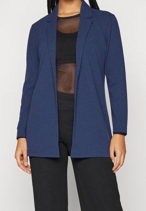 Personne portant un blazer ouvert bleu marine sur un crop top noir transparent et un pantalon noir, debout devant un fond clair uni.