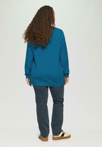 Langsleeve teal sweater gemaakt van zacht materiaal, gecombineerd met donkere denim broek en witte sneakers, die een relaxte pasvorm en casual stijl toont.