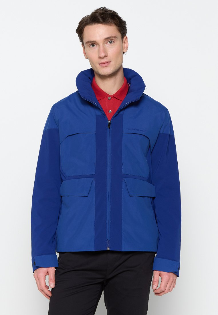 Chaqueta impermeable azul con capucha, que presenta un cierre zipper completo, dos bolsillos frontales con solapa y paneles en contraste en azul más oscuro.