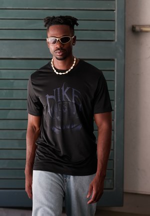 Black Nike graphic t-shirt with blue logo, short sleeves, paired with light blue jeans. Accessorized with shell necklace and sunglasses.
