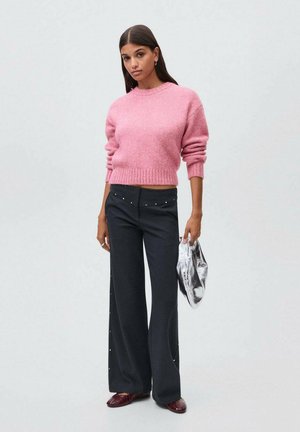 Mujer con un suéter rosa, pantalones anchos negros con pequeños tachuelas, zapatos burdeos, sosteniendo un clutch plateado metálico frente a un fondo simple.
