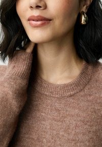 Hellbraunes, gestricktes Sweatshirt mit ribbeligem runden Ausschnitt; kombiniert mit metallischen Tropfenohrringen. Weiche Textur und gemütliches Erscheinungsbild.