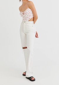 Hvid cropped top med røde prikker, justerbare stropper, parret med højttaljede hvide jeans med et knæudskæring. Sorte slidesandaler.