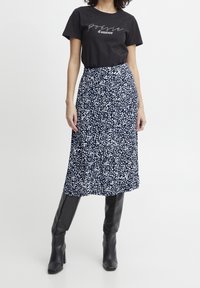 Žena v černém tričku s potiskem "poésie d'amour", námořnických modrých midi sukni s bílým vzorem a černých vysokých kozačkách na podpatku.