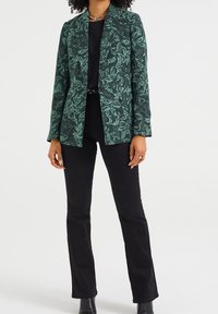 Kvinna klädd i en mörkgrön kavaj med bladmönster över en svart topp, svarta byxor, svarta stövlar och ett gyllene kedjehalsband, stående rakt.