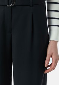 Schwarze Hose mit dezenten Falten, ausgestattet mit einer silbernen Gürtelschnalle. Gestreifter Hemdärmel mit gerippten Bündchen. Glatte Stoffstruktur.