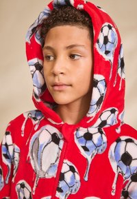 Tuta intera rossa in tessuto morbido con motivo ripetuto di palloni da calcio e dettagli blu. Design con cappuccio, cerniera integrale e texture soffice.