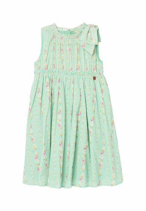 Jeune fille souriante en marchant, portant une robe sans manches bleu clair à motifs floraux et des sandales tressées, avec un fond beige.
