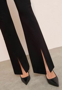 Černé rozšířené kalhoty mají na spodním lemu boční rozparky a jsou vyrobeny z hladkého materiálu. V kombinaci s černými lodičkami na jehlovém podpatku s texturovaným povrchem.