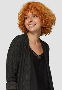 Donkergrijze cardigan met ribbeltextuur, open voorkant en lange mouwen, gedragen over een zwarte top met een kanten halslijn. Krullend oranje haar.