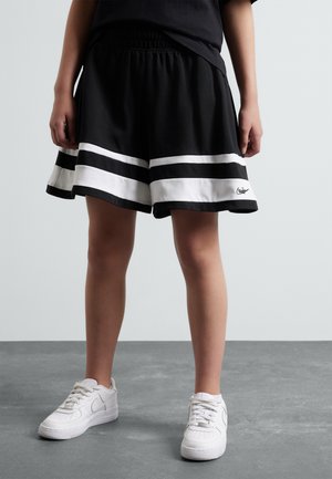 Pantalones cortos deportivos negros con acentos de rayas blancas en el dobladillo, con cinturilla elástica y un parche con el logo en la pierna. Combinados con zapatillas blancas.