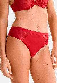 Bas de bikini rouge avec une texture lisse et des panneaux en maille transparente. Dispose d'une taille mi-haute et d'un design simple sans ornements.