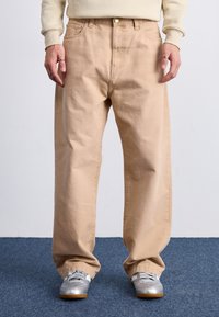 Pantalones de algodón beige con un corte holgado, con cierre de botones y bolsillos. Combinados con zapatos deportivos plateados sobre una alfombra azul.