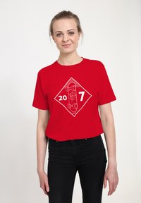 Kvinde med blond hår iført en rød t-shirt med en hvid skateboard og tallene "20" og "7", bærende sorte bukser mod en hvid baggrund.