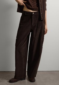 Pantaloni in corduroy a gamba larga di colore marrone scuro con strisce verticali prominenti, chiusura frontale con bottone e tasche laterali. Texture liscia e vestibilità rilassata.