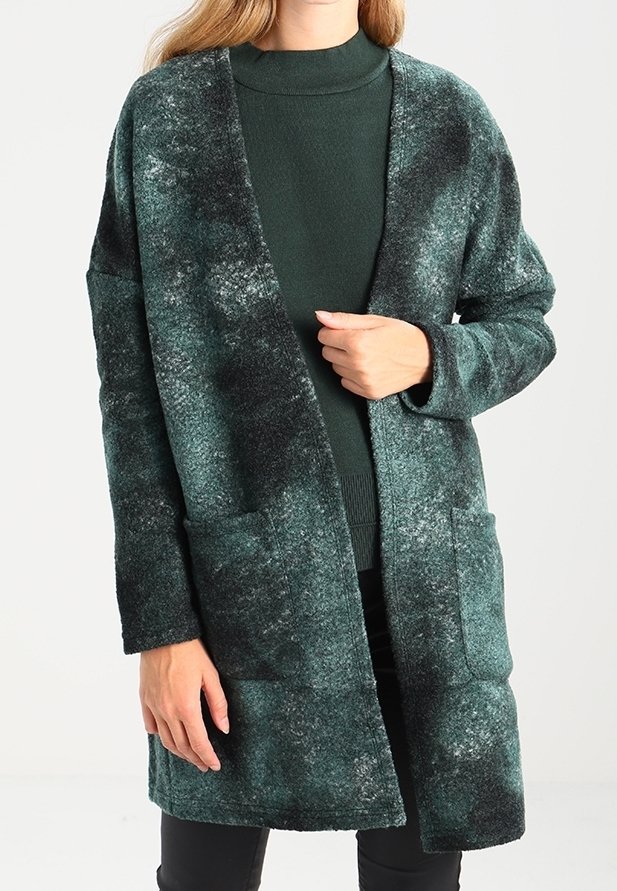 Femme portant un cardigan texturé vert foncé ouvert sur un pull à col roulé vert, avec des poches et des manches longues.