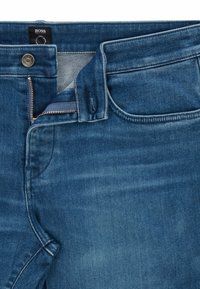 Tmavě modré deniminové džíny se zipem a knoflíkovým zapínáním, s předními kapsami a mírným vyšisováním pro texture. Značková nášivka je viditelná na pase.