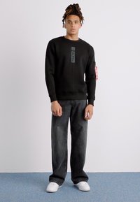 Sudadera negra con logo texturizado, combinada con jeans oscuros de corte suelto y zapatillas blancas. Etiqueta roja notable en la manga.