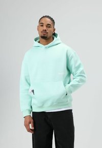 Mintgroene hoodie met een voorzak en capuchon, gemaakt van zacht materiaal; heeft een klein logodetail op de borst en een label op de zak.