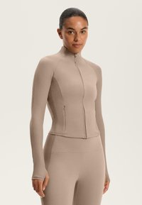 Beige Zip-up Sportjacke mit gerippten langen Ärmeln und körpernahem Schnitt. Glatte Textur mit hohem Kragen und subtilen Details.
