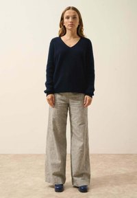 Granatowy sweter w serek z miękkiego materiału, zestawiony z szerokimi spodami w szare paski. Modelka nosi niebieskie buty, stojąc na jednolitym tle.