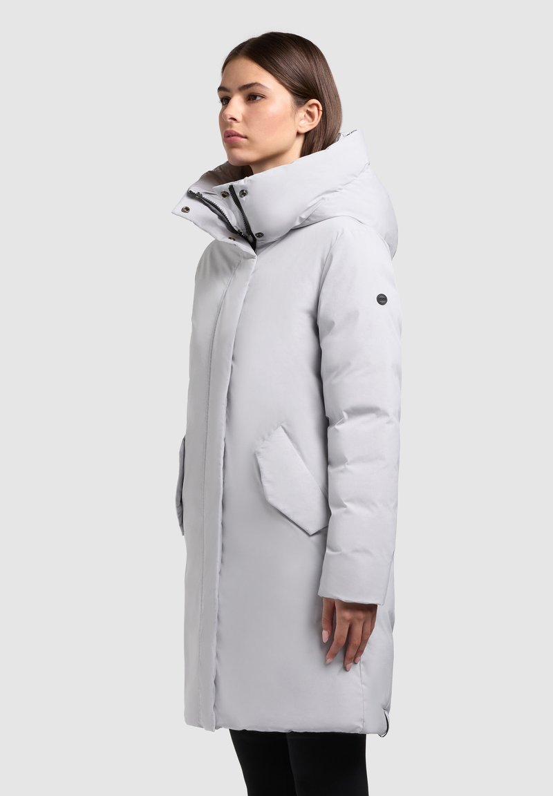 Gaiši pelēks pūkains mētelis ar augstu apkakli, sānu kabatām un rāvējslēdzēja aizdari. Gluda tekstūra un izcelta šuvju detaļa.