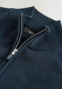 Dark blue knit sweater with a ribbed high collar and a partially open silver zipper showing the NEXT brand label inside.