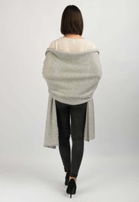Grå sjals-agtig omslag, blød uldblanding, der hænger over skuldrene, med længere bagpaneler, båret med mørke jeans og høje hæle.