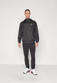 Svart lättviktsvindjacka med dragkedja fram och elastiska ärmslut, ihop med svarta joggingbyxor som har en vit logga. Vita skor med röda detaljer.