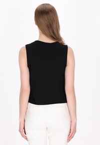 Top nero senza maniche con scollo rotondo, design leggermente corto e texture liscia. Indossato con pantaloni di colore chiaro.