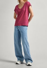 T-shirt a maniche corte, scollo a V profondo, color bordeaux abbinato a jeans a gamba larga blu chiaro. Sneakers bianche completano l'outfit. Sfondo neutro.