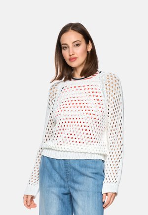 Frau trägt weißen, locker gestrickten Pullover über einem rot-weiß gestreiften Oberteil und hellblauen Jeans, steht vor einfarbigem Hintergrund.