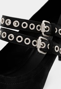 Μαύρα σουέντ τακούνια με δύο ρυθμιζόμενες λουρίδες με ασημένια τρύπες και αγκράφες, προσθέτοντας ένα διακριτικό σχεδιαστικό στοιχείο στα παπούτσια.