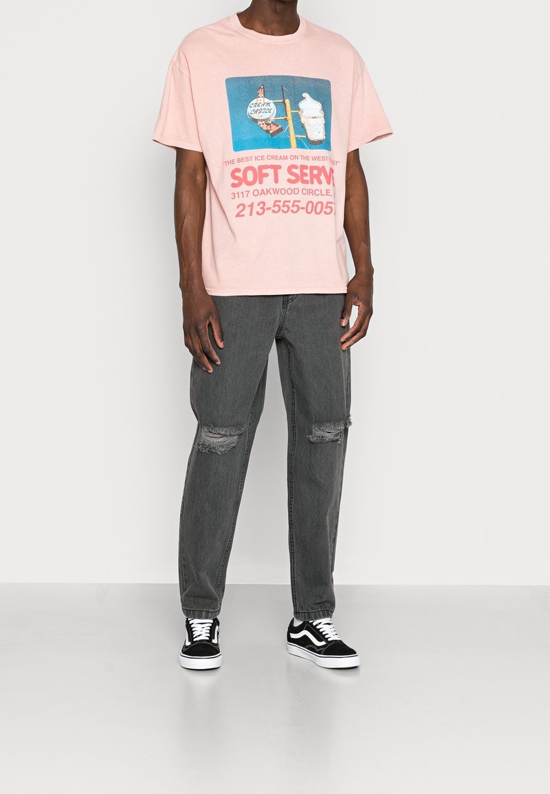 Muž v růžovém grafikou potištěném tričku s reklamou na zmrzlinu, v roztrhaných tmavě šedých džínách a černobílých teniskách stojící na jednobarevném pozadí.