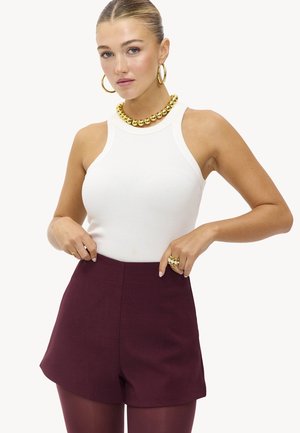 Débardeur blanc à côtes, short taille haute bordeaux, collier chunky doré et boucles d'oreilles. Le mannequin pose les mains sur les hanches.
