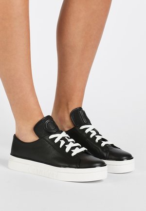 Feet wearing black leather sneakers with white laces and thick white soles, standing on a plain light background.