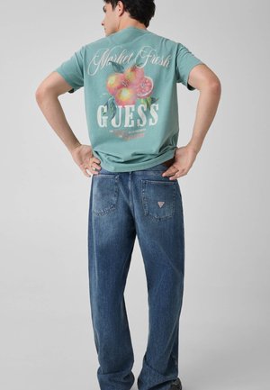 Homme portant un t-shirt turquoise avec un motif pêche et le texte « "Market Fresh Guess" », associé à un jean bleu, debout, mains sur les hanches, dos tourné.