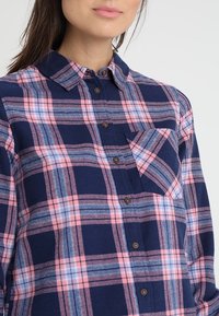 Blue and pink plaid button-up shirt with a collar, long sleeves, and a chest pocket. Features brown buttons and a textured fabric.