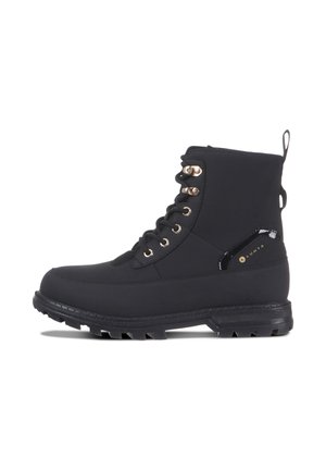 Bota negra de caña alta con cordones, ojales metálicos y suela gruesa con tacos, con lengüeta para tirar y detalle lateral con marca.