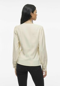 Blusa da donna in crema con maniche lunghe, scollo rotondo e spalle leggermente a palloncino; presenta dettagli con bottoni ai polsini.