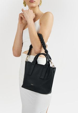 Donna in abito bianco che tiene una borsa nera in pelle con tracolla intrecciata e dettagli dorati su sfondo uniforme.