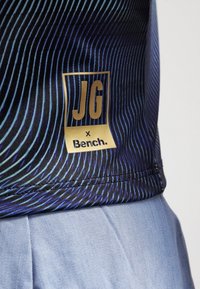 Gros plan sur l'ourlet d'une chemise rayée bleue et noire avec une étiquette dorée indiquant "JG x Bench." sur un pantalon bleu clair.