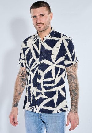 Man with short hair and tattoos on both arms wearing a short-sleeve navy and white abstract patterned shirt and light blue jeans.