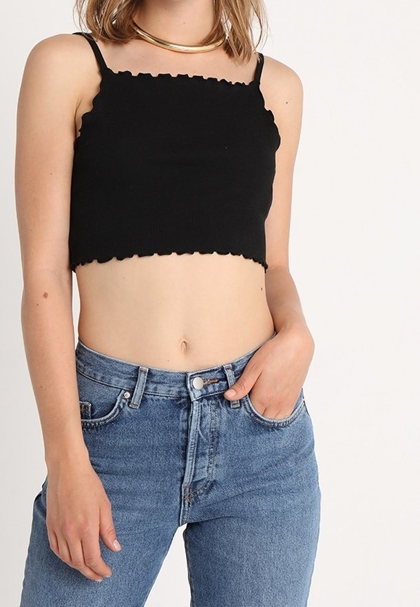 Femme portant un crop top noir à fines bretelles et un jean bleu taille haute, main dans la poche, sur un fond uni.