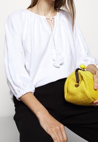 Biała, teksturowana bluzka z bufiastymi rękawami i zapięciem na kokardkę, w połączeniu z małą, okrągłą, żółtą torbą plecioną z czarnym zapięciem i żółtym akcentem.